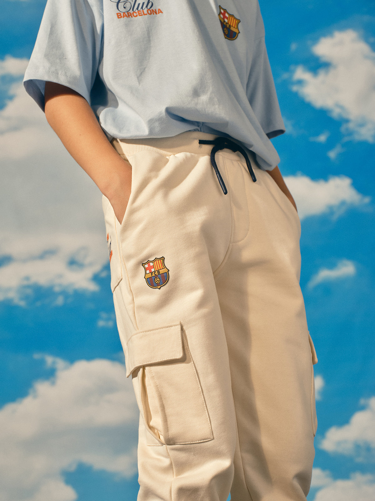 Beige FC Barcelona kids' jogger pants with side cargo pockets, elastic cuffs, drawstring waist, and club crest on the upper leg.