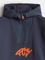 Gros plan d’une veste à capuche bleu marine FC Barcelona avec col montant zippé et logo orange Barça sur la poitrine.