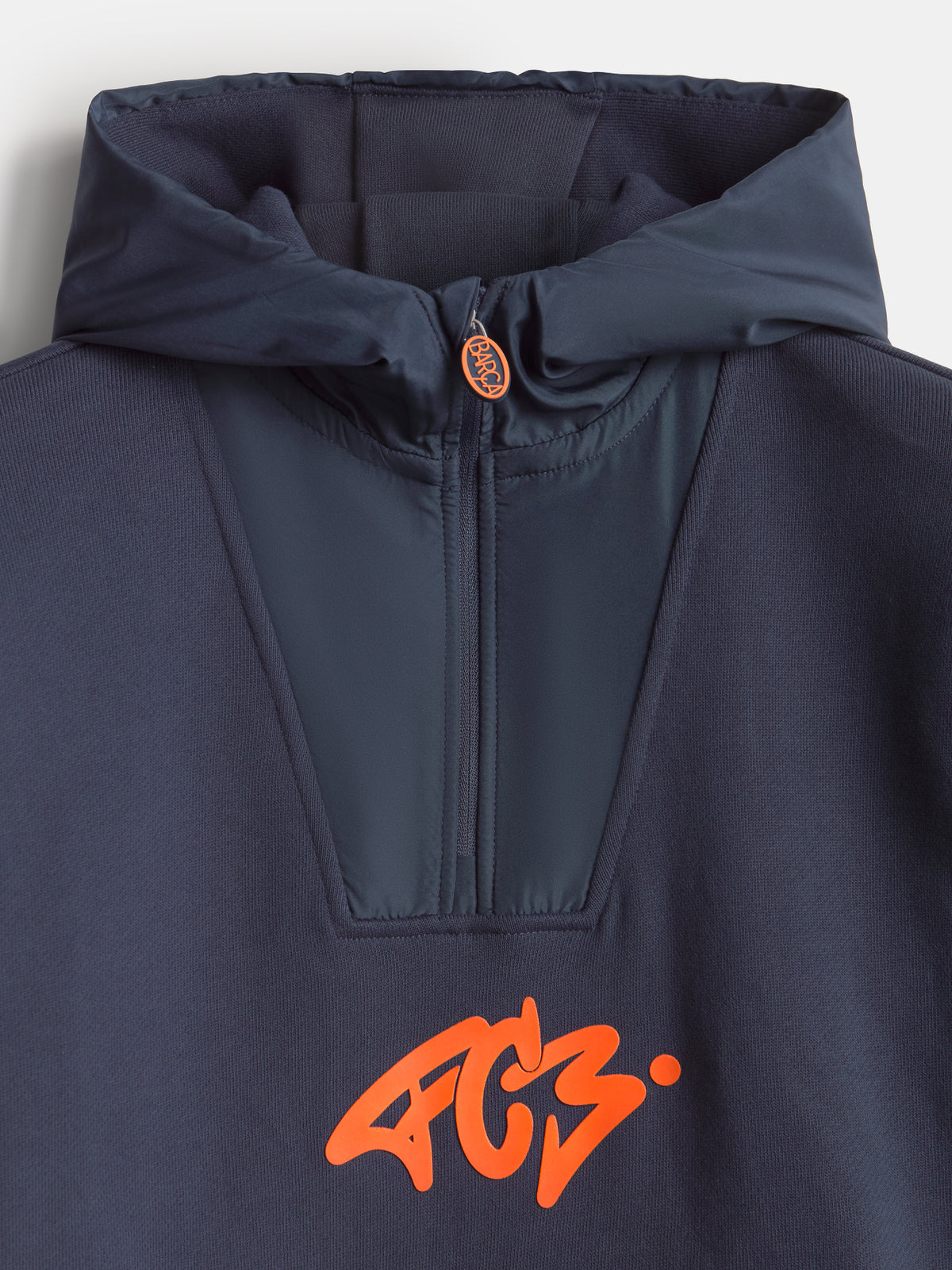 Gros plan d’une veste à capuche bleu marine FC Barcelona avec col montant zippé et logo orange Barça sur la poitrine.