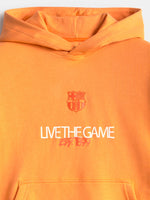 Vista frontal de una sudadera con capucha naranja de FC Barcelona con el escudo del club y el texto “LIVE THE GAME” en el pecho.