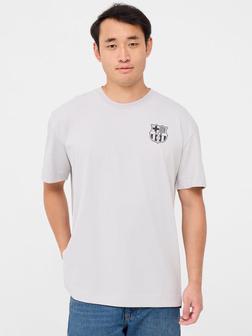 Back of a light beige T-shirt featuring a playful blue FC Barcelona crest cartoon kicking a soccer ball. Above it reads “FC BARCELONA” and below, in bold, “BLAU GRANA SÓN ELS COLORS” with “EST. 1899” on each side
