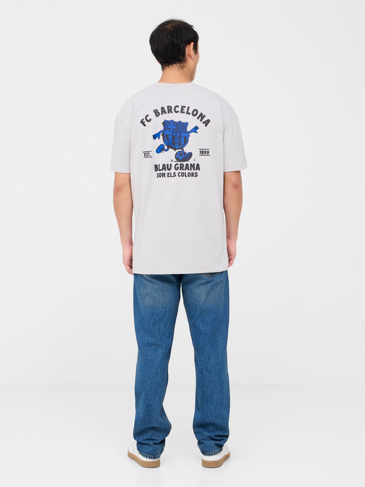 Back of a light beige T-shirt featuring a playful blue FC Barcelona crest cartoon kicking a soccer ball. Above it reads “FC BARCELONA” and below, in bold, “BLAU GRANA SÓN ELS COLORS” with “EST. 1899” on each side