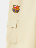 Beige FC Barcelona kids' jogger pants with side cargo pockets, elastic cuffs, drawstring waist, and club crest on the upper leg.