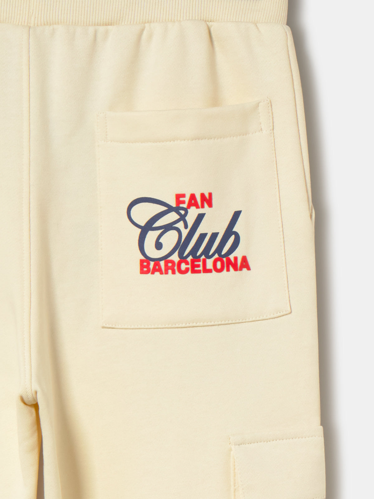 Beige FC Barcelona kids' jogger pants with side cargo pockets, elastic cuffs, drawstring waist, and club crest on the upper leg.