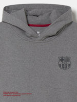 Front view of a grey FC Barcelona hoodie with a cowl-style hood, dark Barça crest on the chest, and small red text near the hem.