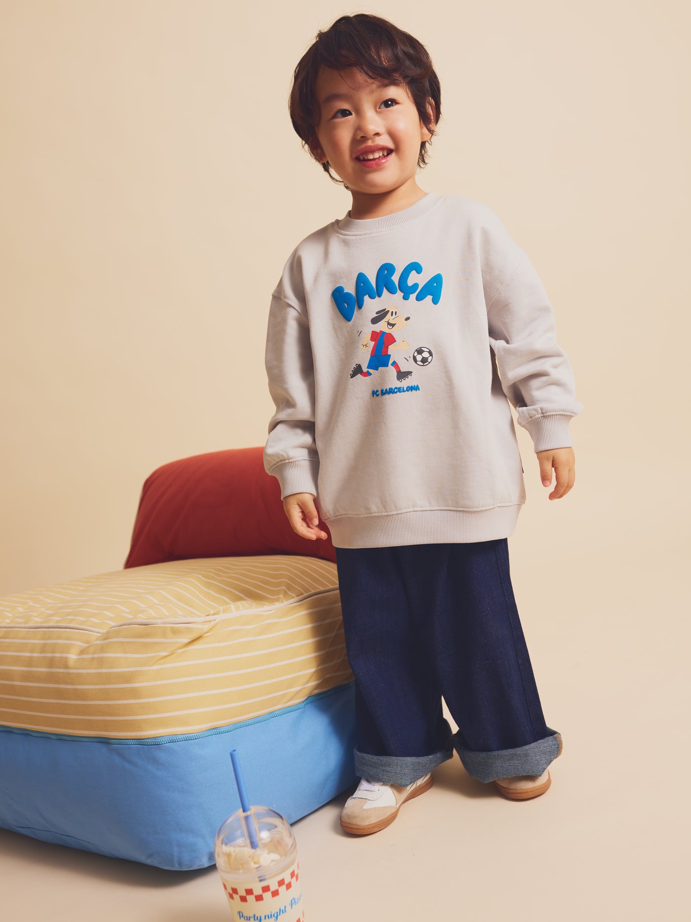 Smiling child standing next to a cushion, wearing a light grey FC Barcelona sweatshirt with a cartoon footballer and the word 'Barça' in blue