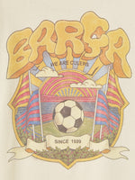 Beige FC Barcelona T-shirt featuring a vibrant vintage-style back print with the word "Barça" in large retro bubble letters. The graphic includes a football, flags, and stadium elements in bold colors within a shield-like frame. A ribbon below the design reads “Since 1899” and “Més que un club.”