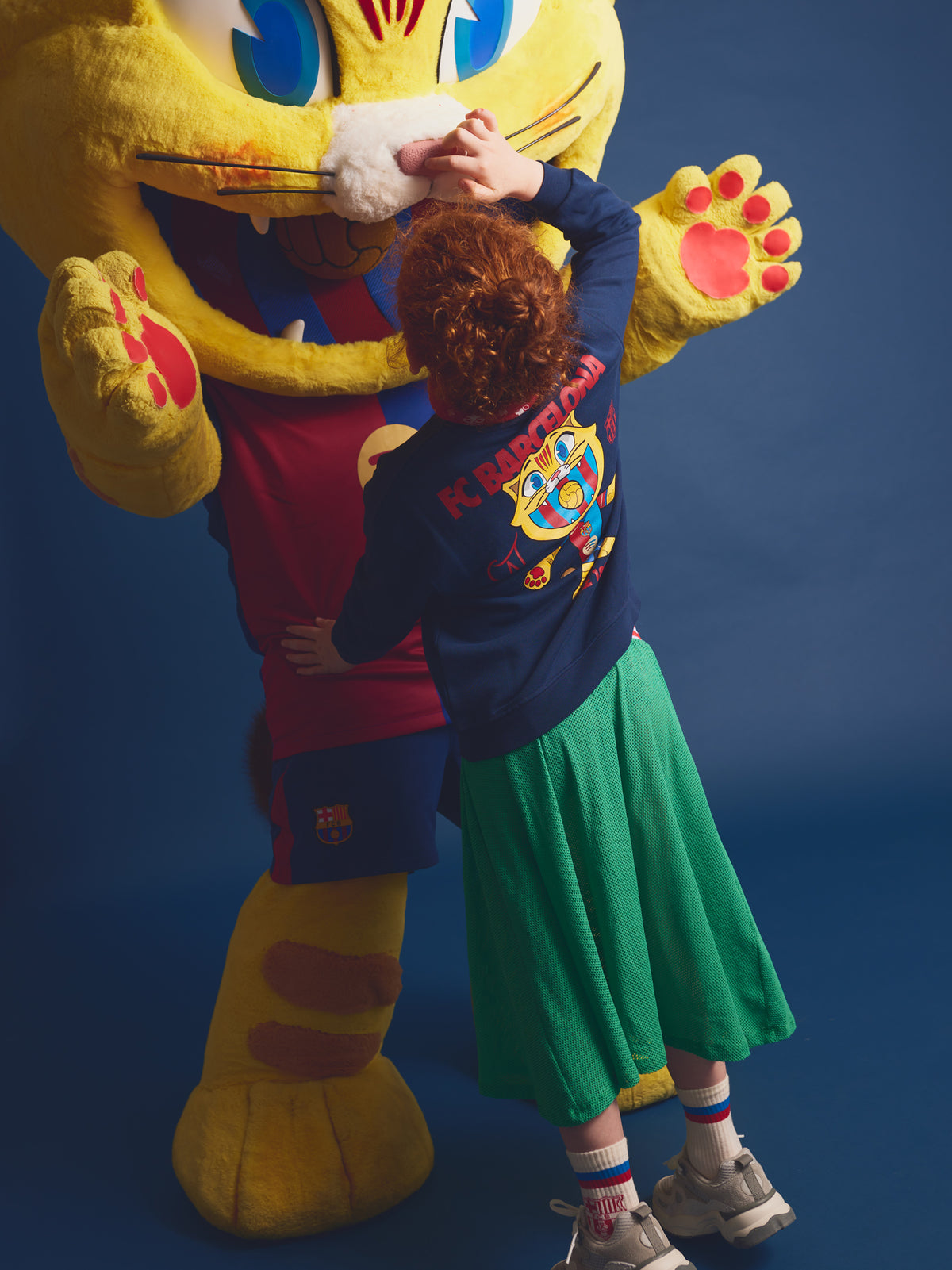 Girl hugging Barça’s mascot, Cat Culer, wearing a navy blue sweatshirt with a colorful Cat Culer illustration on the back, green skirt, and Barça socks.