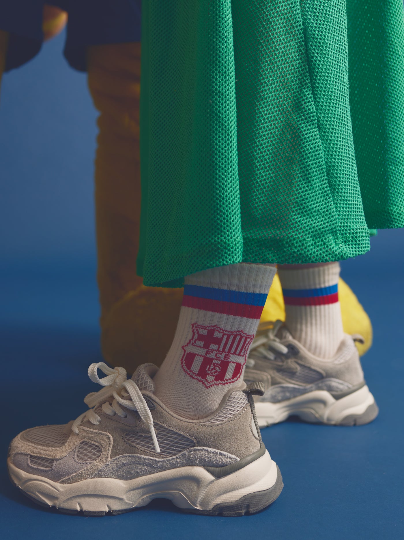 Close-up of a child wearing white FC Barcelona socks with red and blue stripes and the club crest, paired with grey sneakers and green mesh pants.