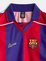 Vista frontal de una camiseta del FC Barcelona con franjas verticales rojas y azules, cuello morado y el escudo del club en el pecho.