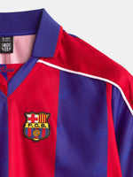 Primer plano de un polo de FC Barcelona con rayas verticales rojas y azules, cuello morado y escudo del club en el pecho.
