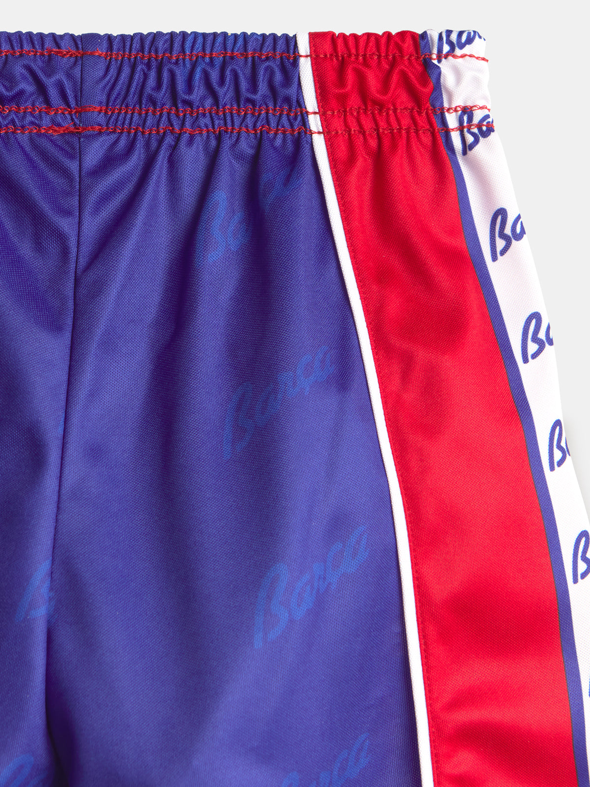 Gros plan sur un short FC Barcelona à base violette, panneau latéral rouge et bordure blanche avec imprimé répété du logo Barça.