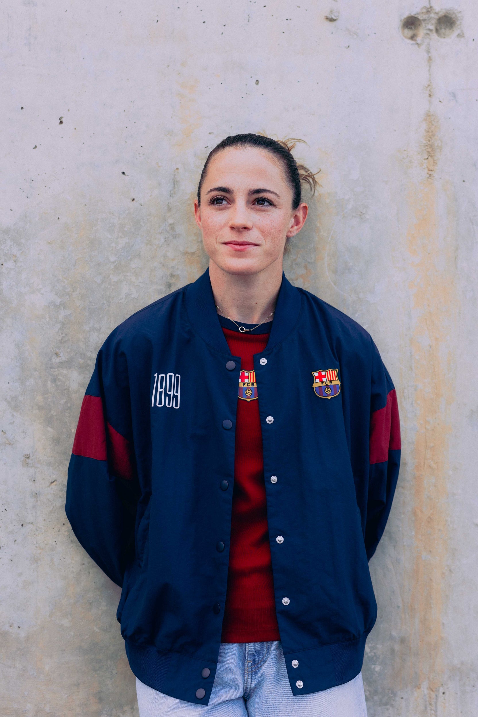 Person wearing a blue jacket with red accents and logos, standing against a textured wall.