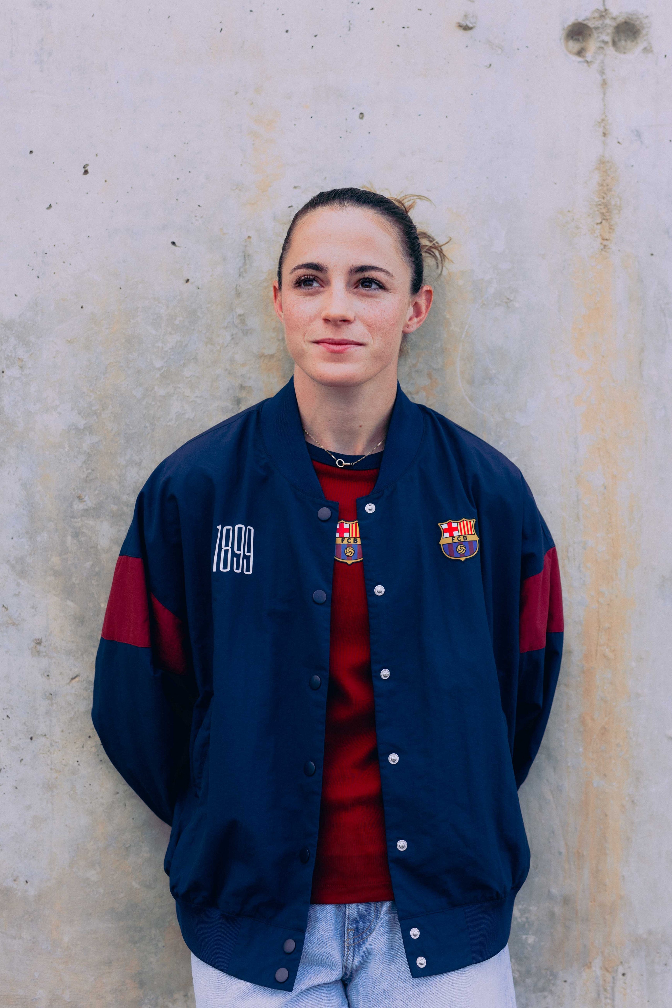 Person wearing a blue jacket with red accents and logos, standing against a textured wall.