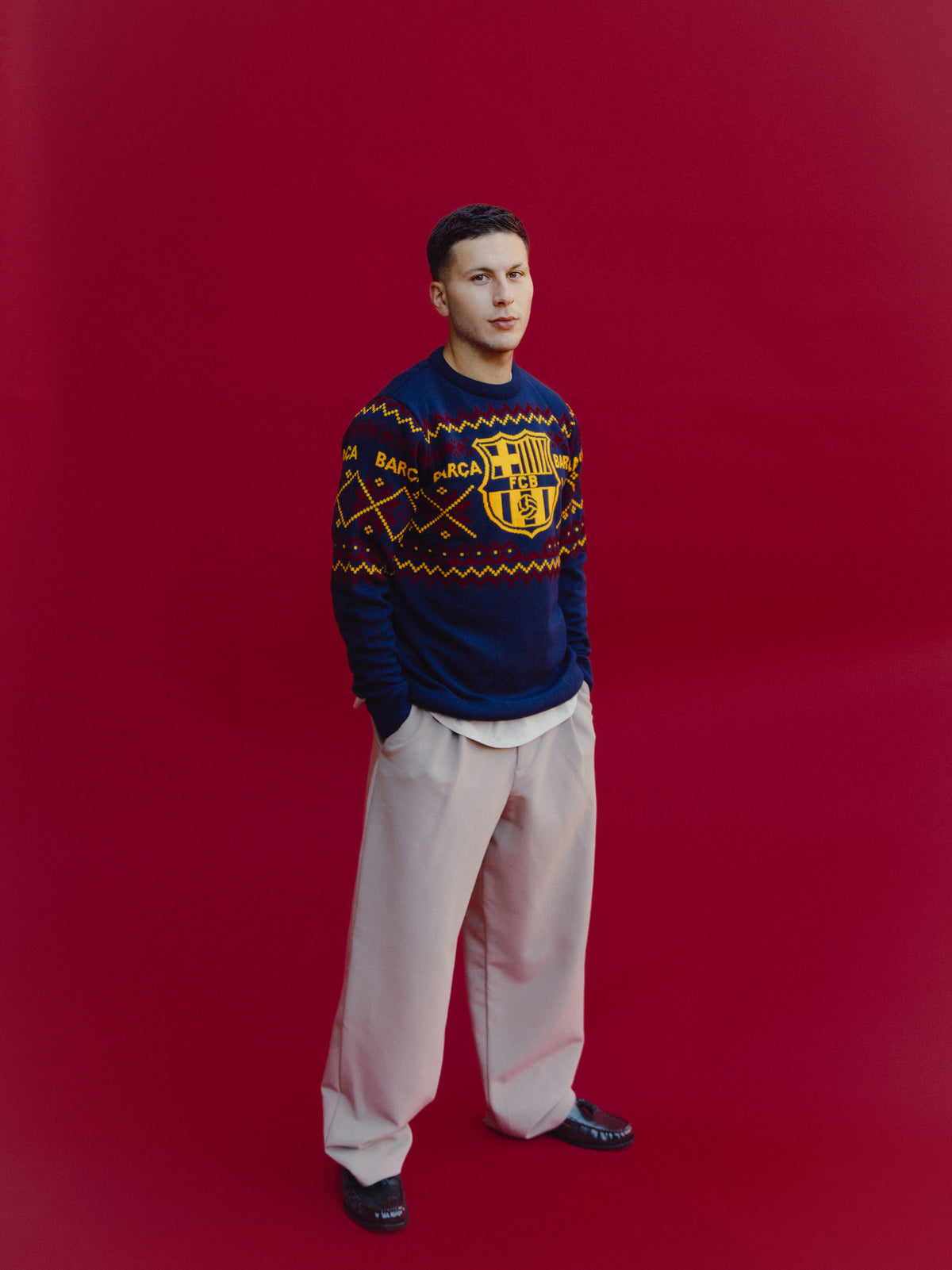 Mannequin en pied portant un pull FC Barcelona bleu marine avec un grand écusson jaune et une bande à motif sur la poitrine, avec un pantalon beige.