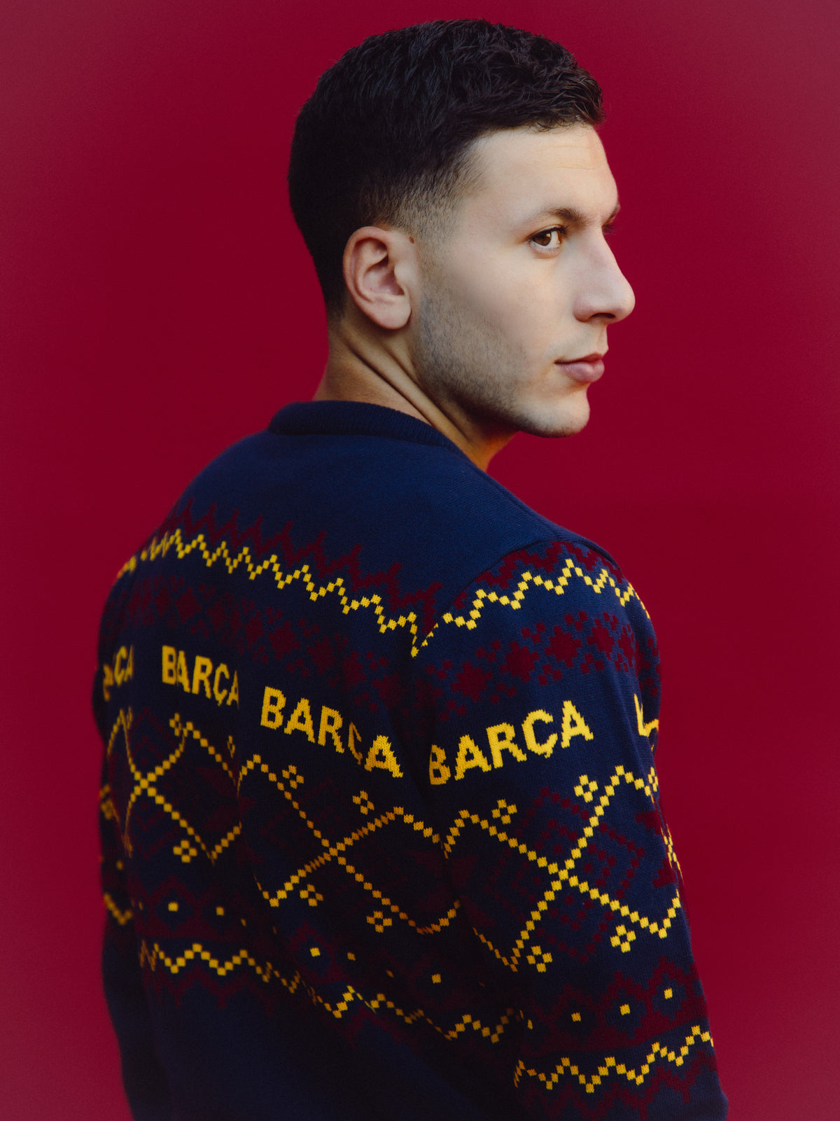 Modèle portant un pull bleu marine FC Barcelona avec lettrage BARCA jaune et motif géométrique, vu de profil sur fond rouge