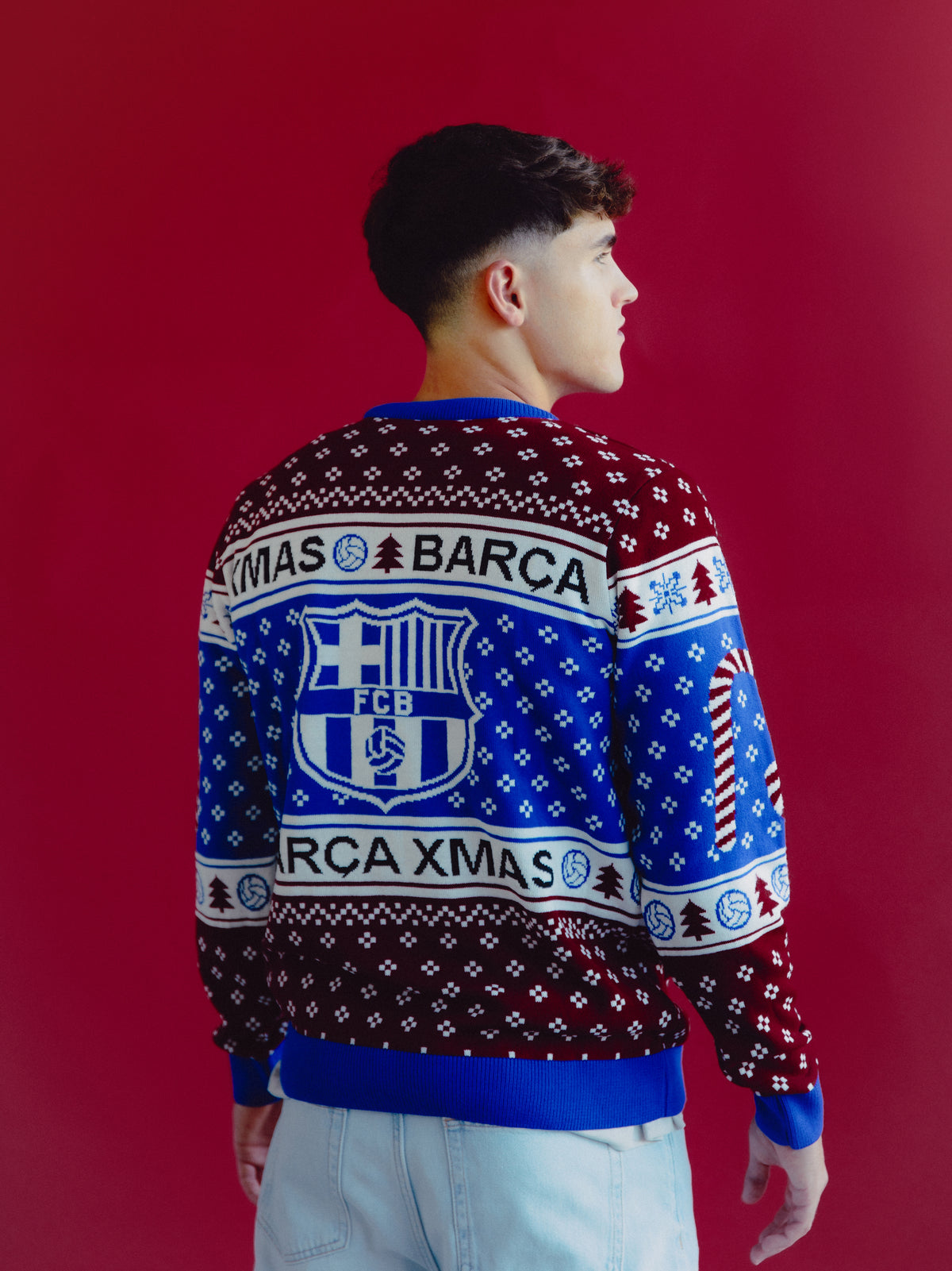 Vista trasera de un modelo con un jersey festivo de FC Barcelona con un gran escudo FCB y el texto «XMAS BARÇA» sobre un patrón rojo, azul y blanco.