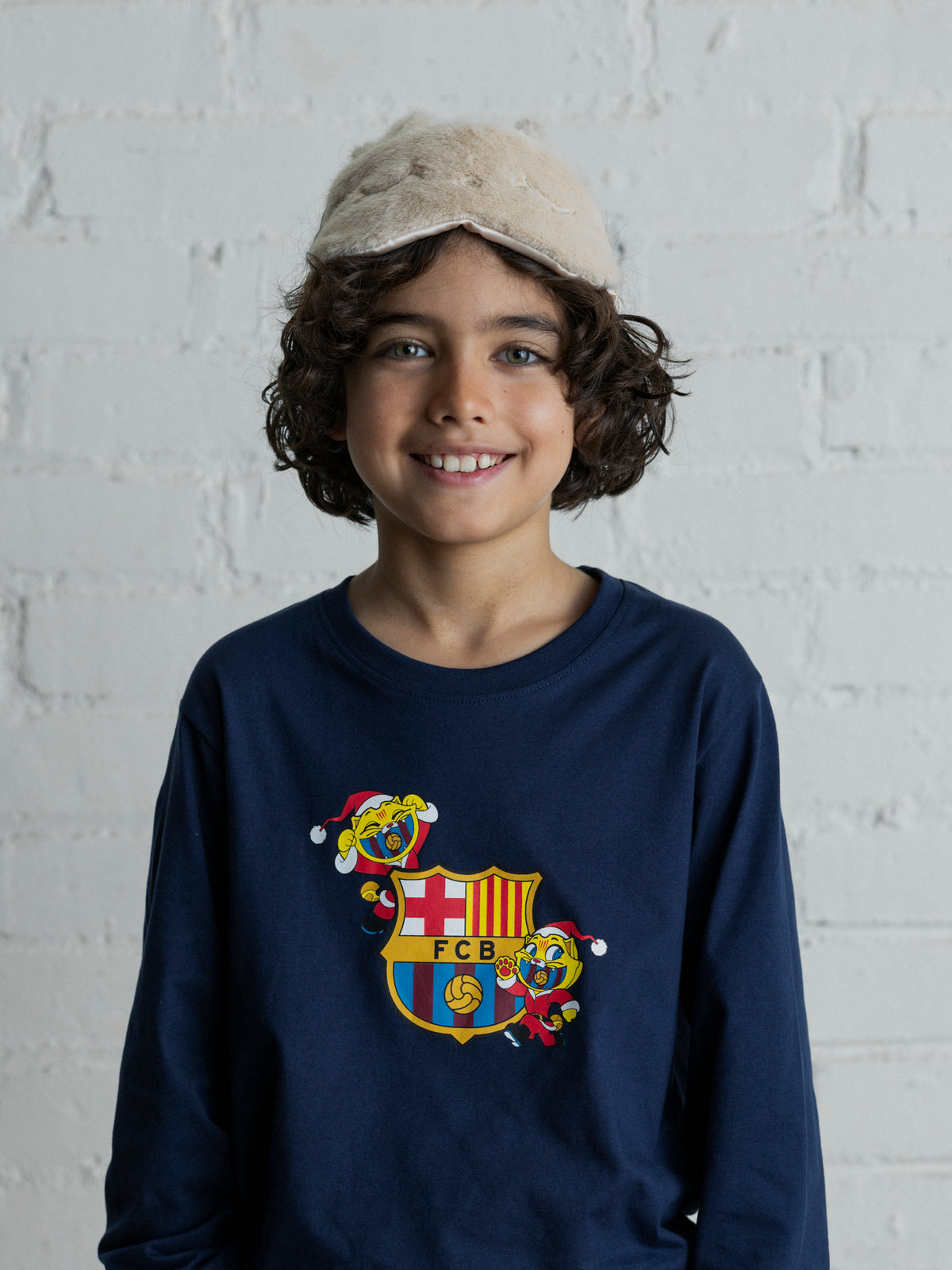 Enfant portant un maillot FC Barcelona bleu marine à manches longues, avec un motif festif de l’écusson sur le devant et un bonnet duveteux beige.