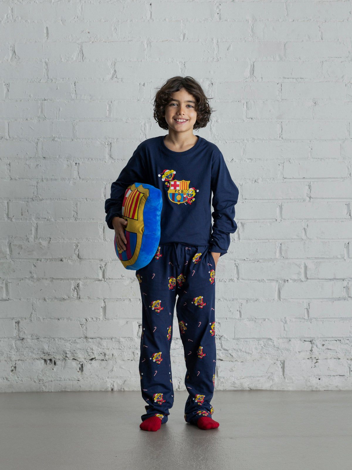 Enfant portant un pyjama bleu marine du FC Barcelona avec imprimé coloré du blason de Barça, tenant un coussin bleu et rouge du FC Barcelona.