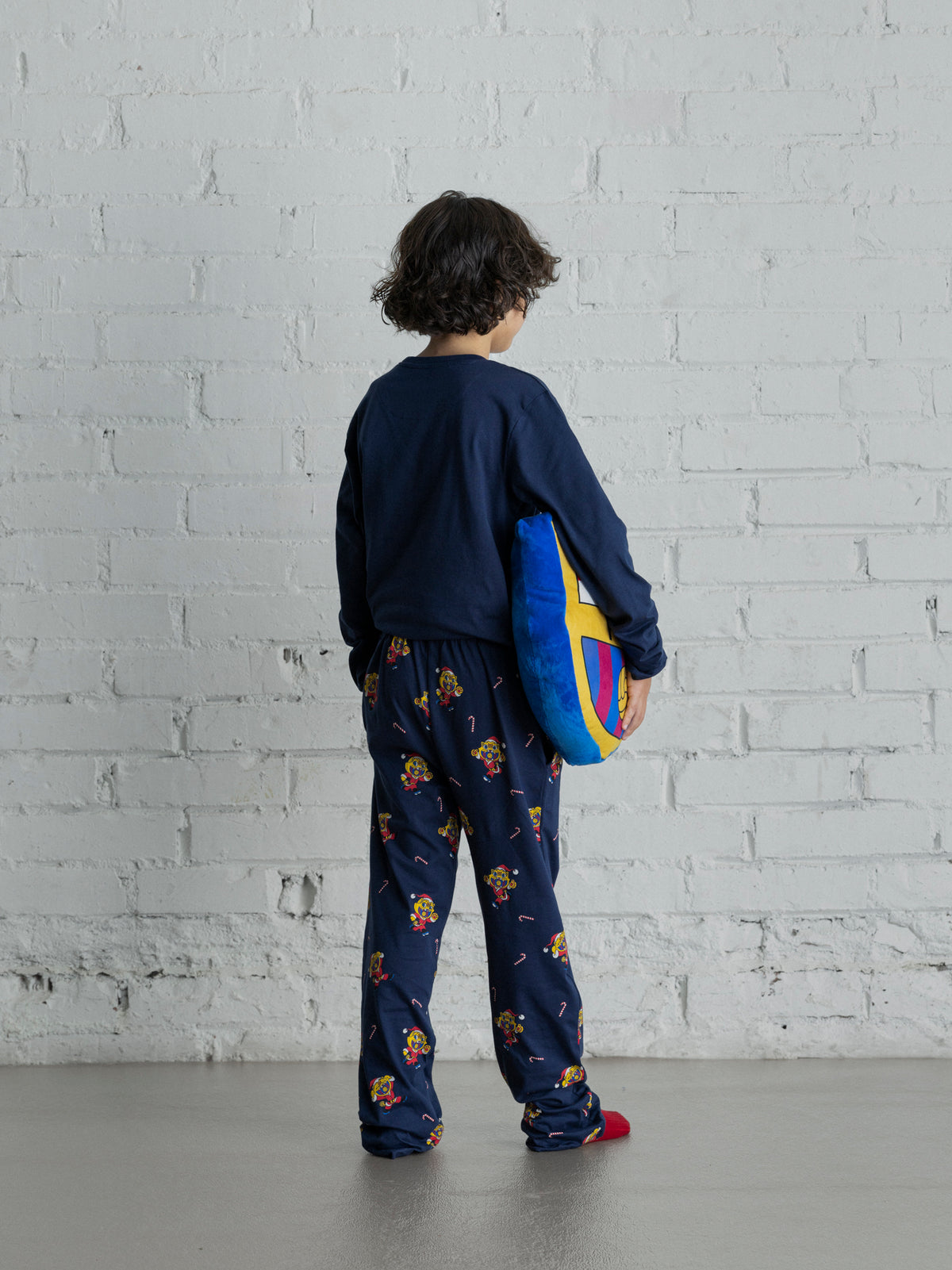 Vue arrière d’un enfant portant un pyjama bleu marine FC Barcelona avec des motifs de dessins animés colorés et tenant un coussin Barça bleu, jaune et rouge.