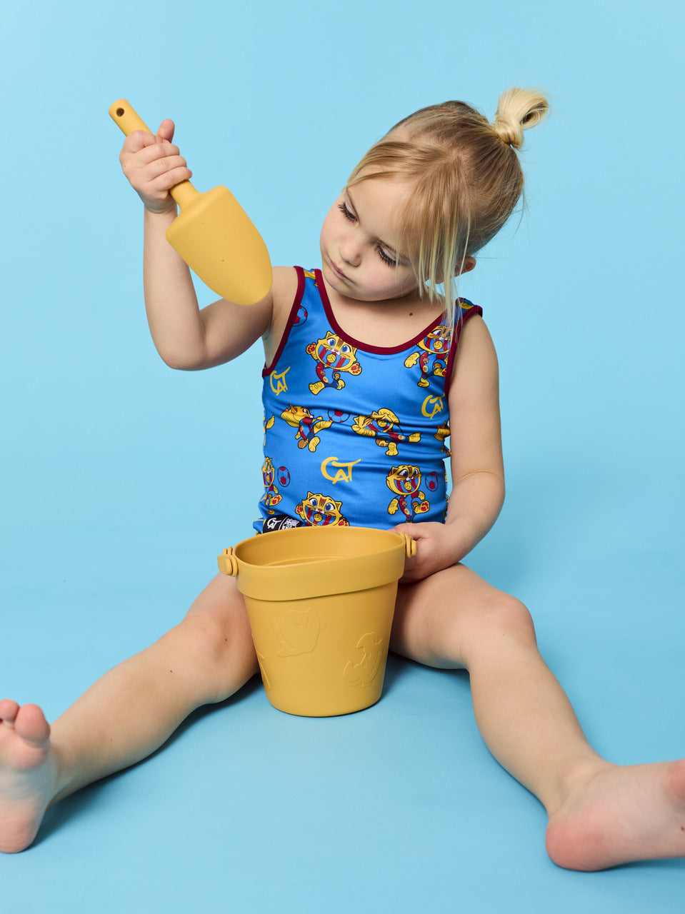 Child in a blue swimsuit with cartoon characters, holding a yellow shovel and bucket on a blue background
