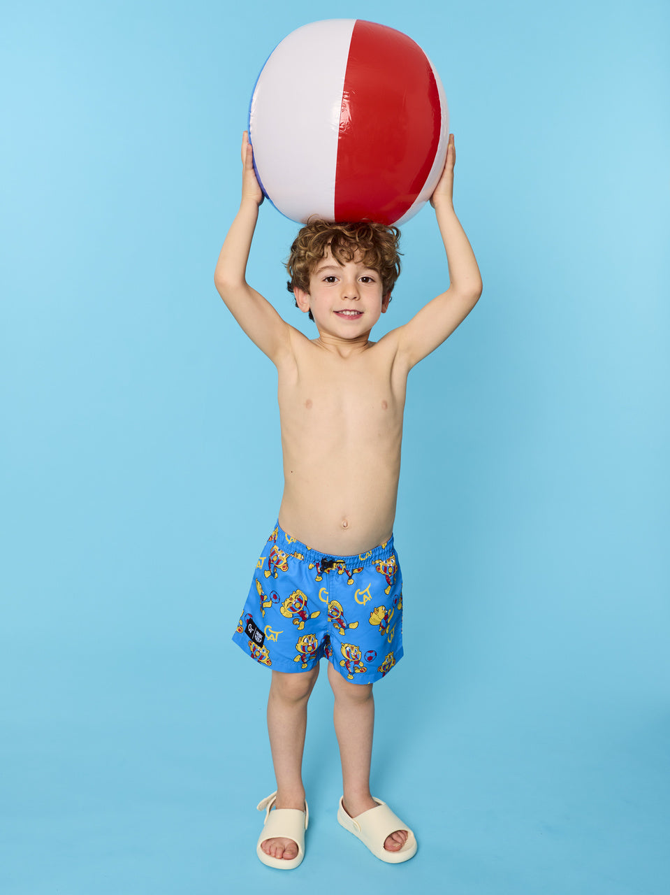 Child wearing blue swim trunks with a red, white, and blue beach ball on a light blue background
