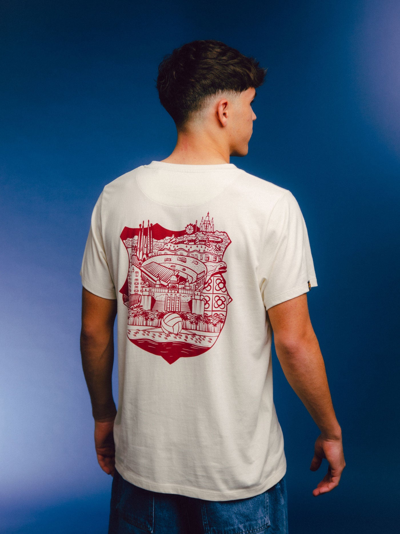 Man wearing an off-white FC Barcelona t-shirt with a red artistic illustration on the back, featuring iconic landmarks of Barcelona and the Camp Nou stadium inside a stylized club crest.