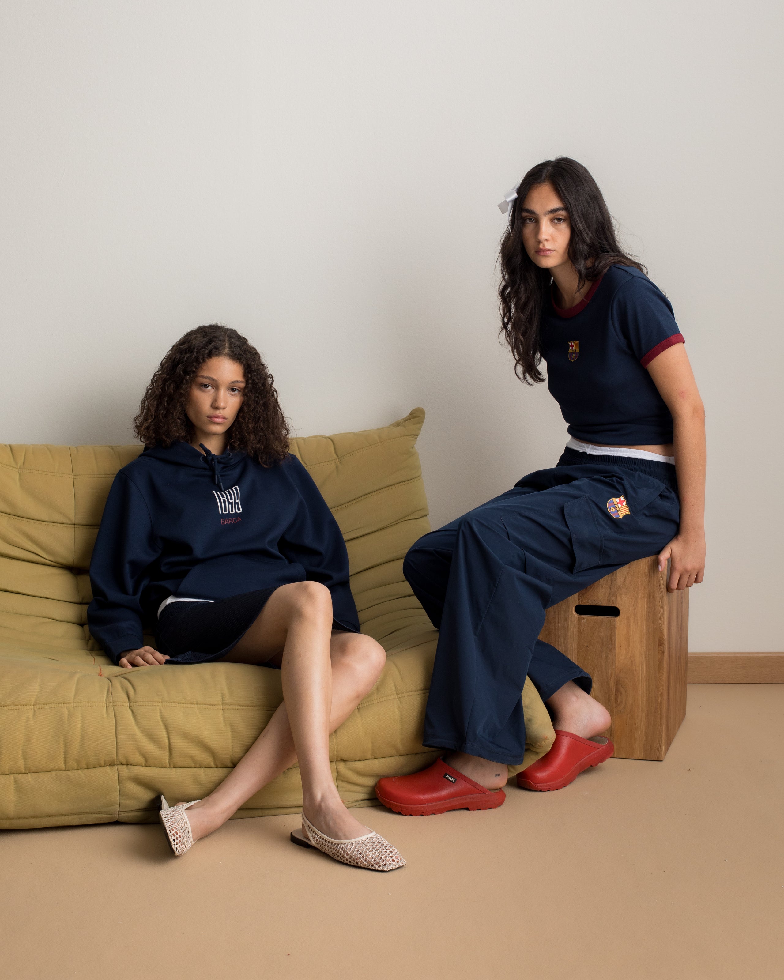Two women wearing FC Barcelona apparel: one in a navy hoodie with white text and a black skirt, the other in a navy crop top with crest and wide-leg pants, sitting indoors on a sofa and wooden stool