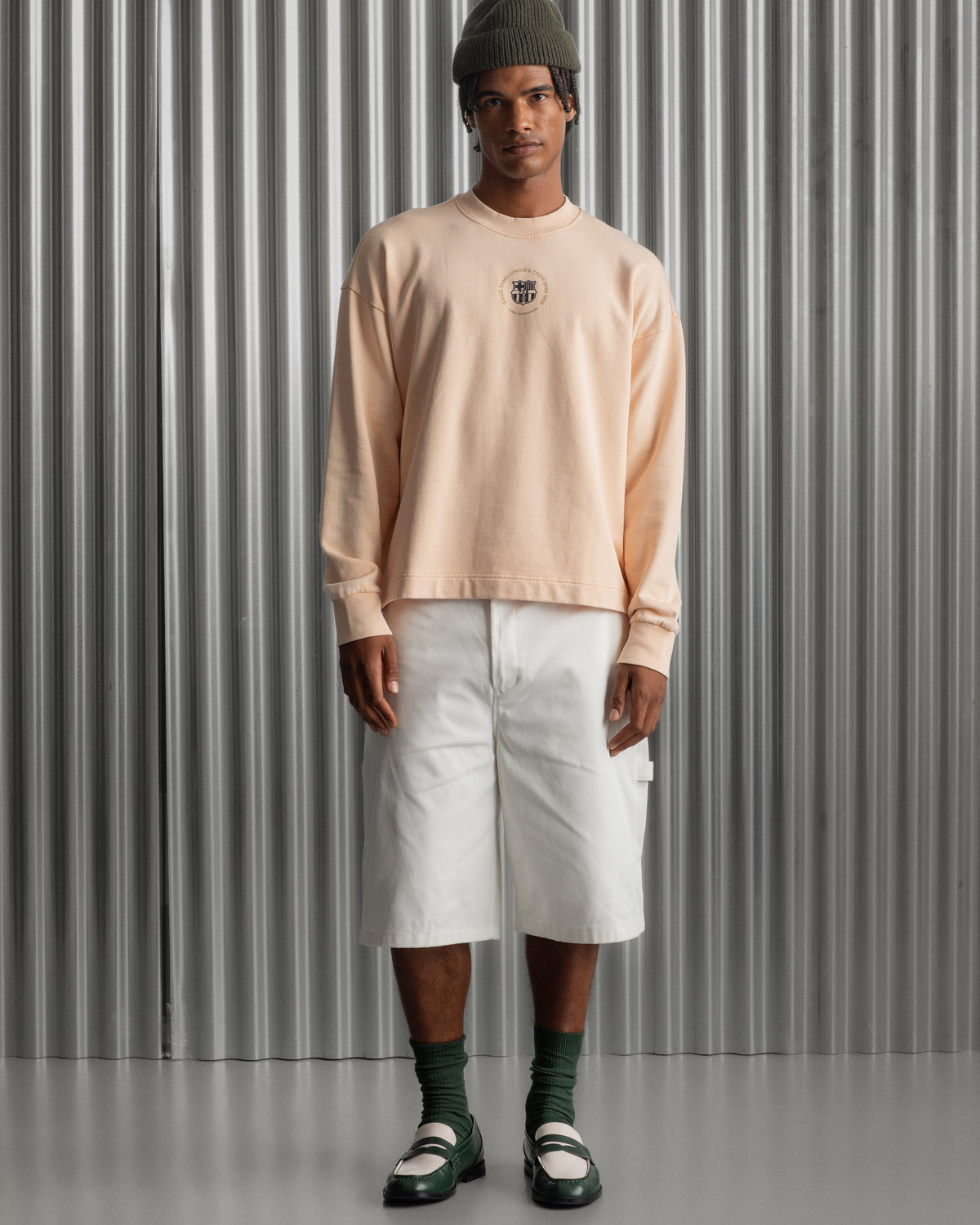 Man wearing a light peach long-sleeve FC Barcelona sweatshirt with a small circular crest detail on the chest, paired with white pants, standing against a metallic background.