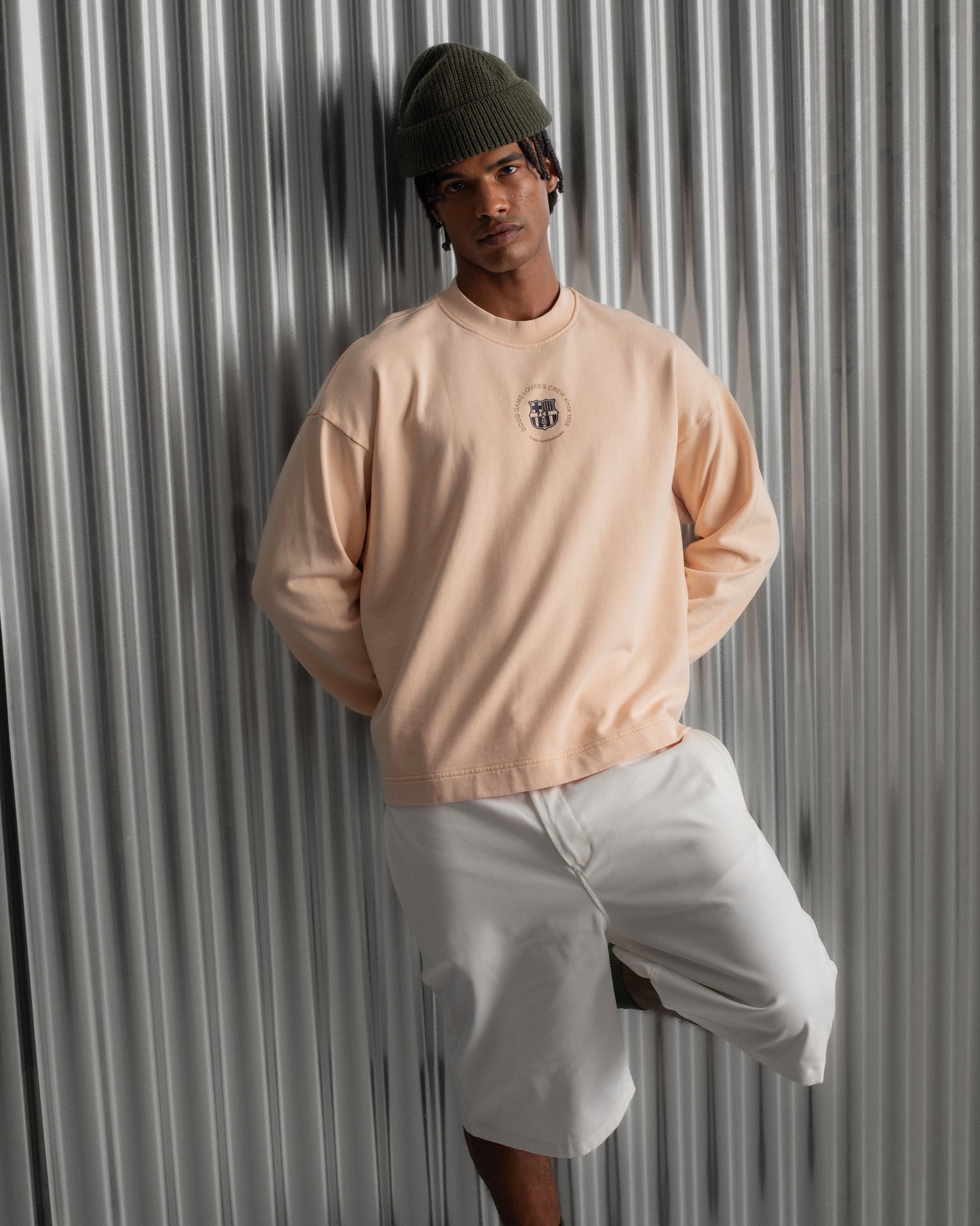 Man wearing a light peach long-sleeve FC Barcelona sweatshirt with a small circular crest detail on the chest, paired with white pants, standing against a metallic background.