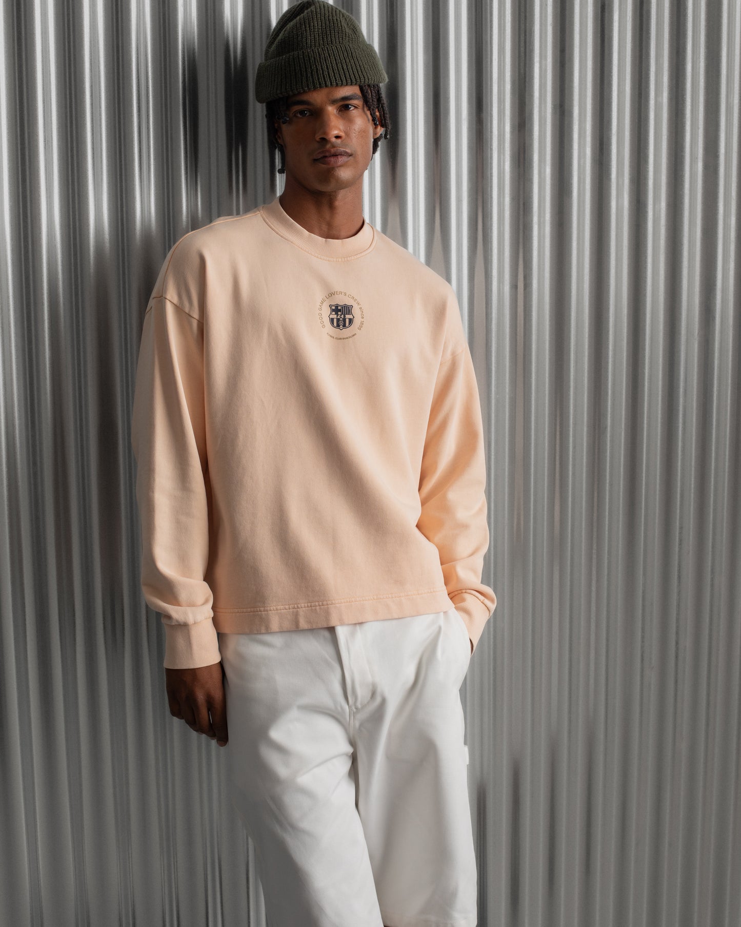 Man wearing a light peach long-sleeve FC Barcelona sweatshirt with a small circular crest detail on the chest, paired with white pants, standing against a metallic background.