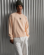 Man wearing a light peach long-sleeve FC Barcelona sweatshirt with a small circular crest detail on the chest, paired with white pants, standing against a metallic background.