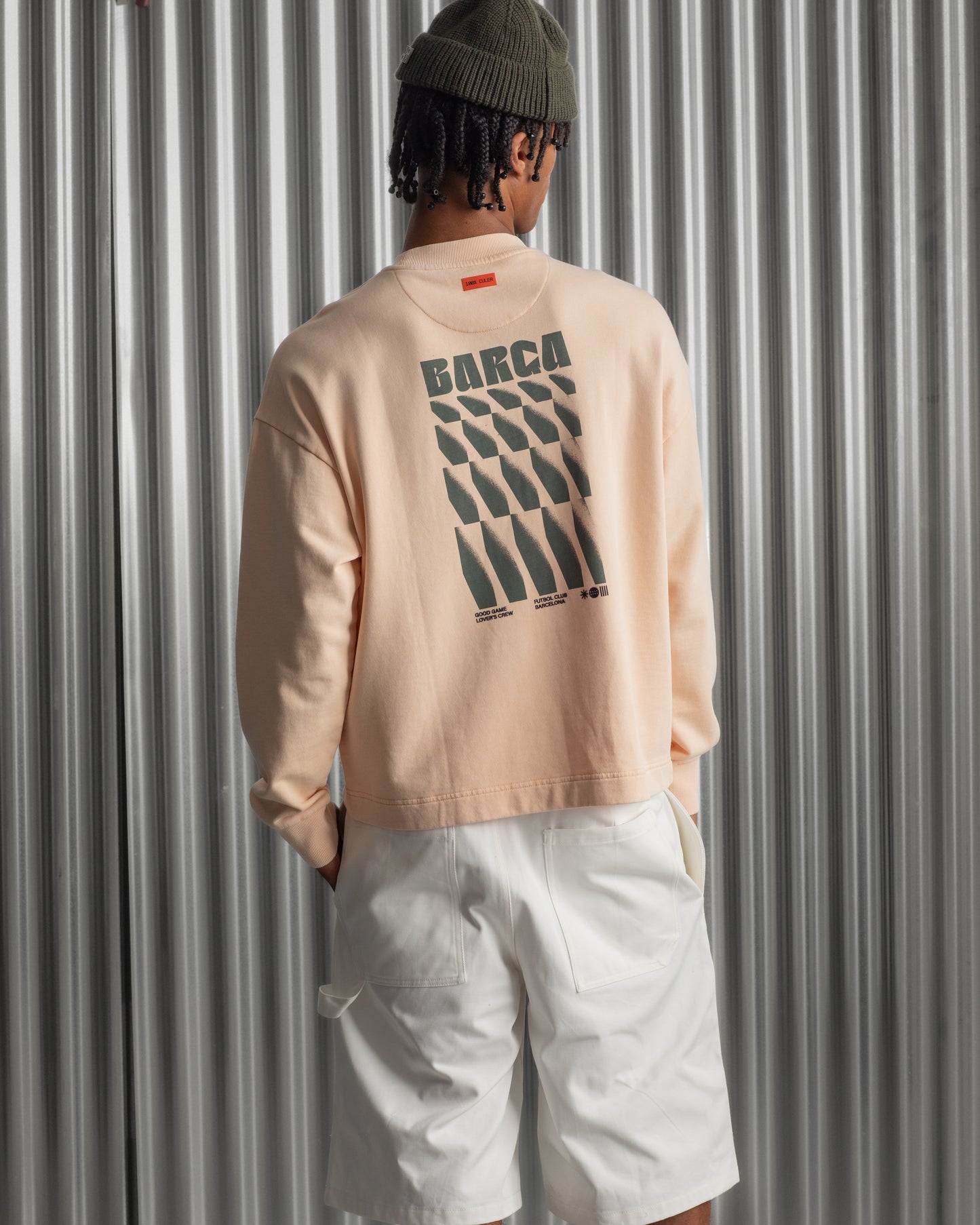 Man wearing a light peach long-sleeve FC Barcelona sweatshirt with a small circular crest detail on the chest, paired with white pants, standing against a metallic background.