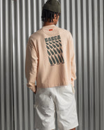 Man wearing a light peach long-sleeve FC Barcelona sweatshirt with a small circular crest detail on the chest, paired with white pants, standing against a metallic background.