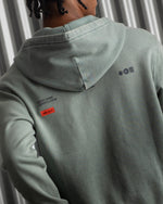 light green FC Barcelona zip-up hoodie with the club’s crest in dark print on the chest, standing against a metallic background.