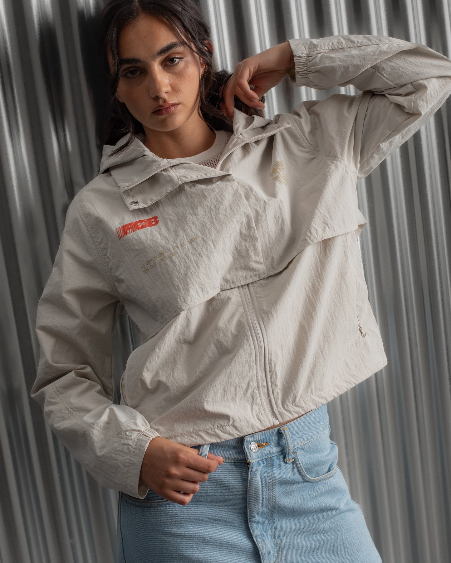 Chaqueta corta beige con capucha del FC Barcelona, cresta bordada en el pecho y detalles gráficos en rojo y dorado, combinada con pantalón vaquero azul claro