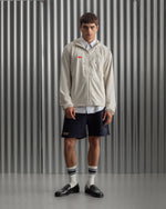 Man wearing a light beige FC Barcelona hooded jacket with front zipper, paired with navy shorts, white shirt, and striped socks, standing against a metallic background.