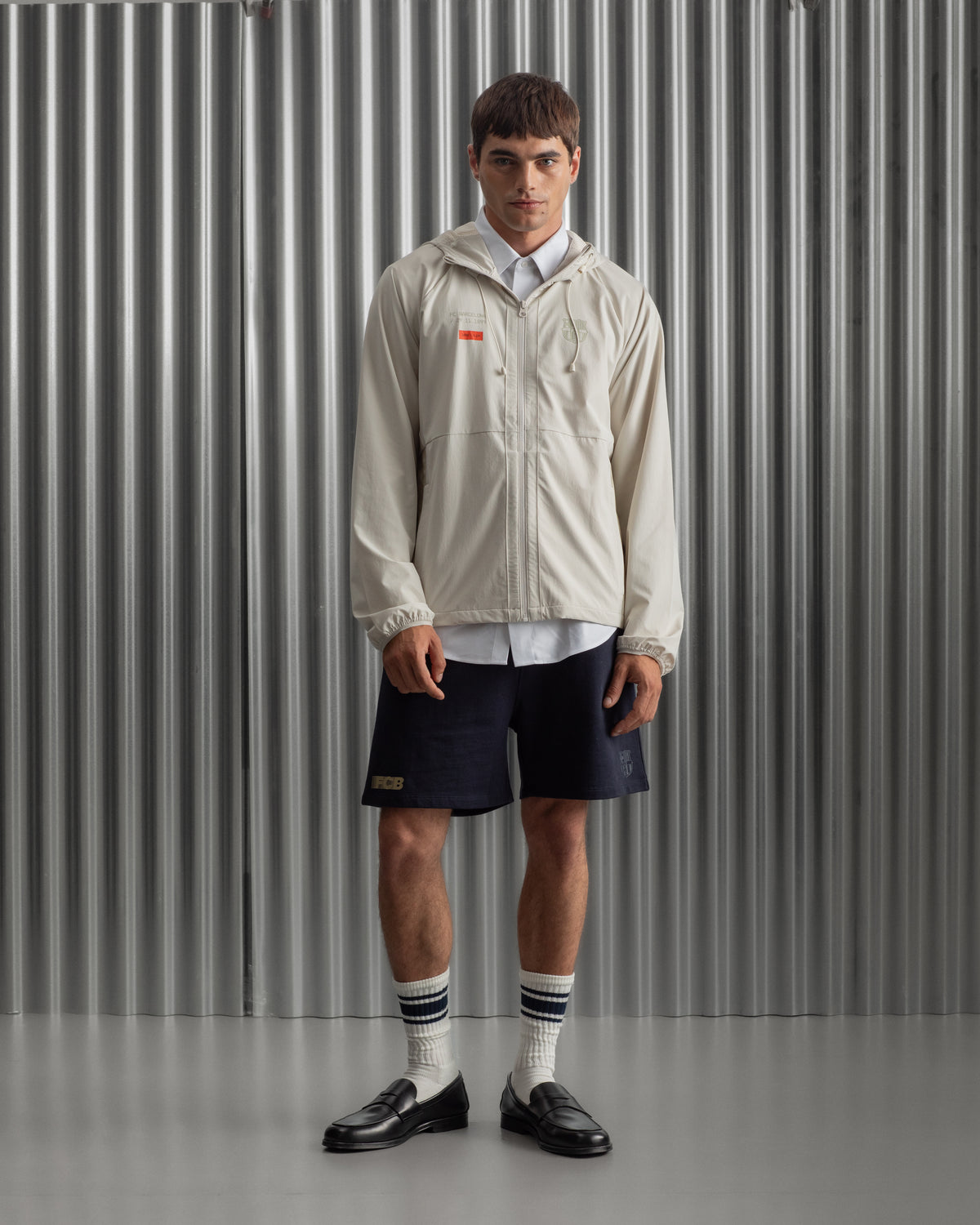 Man wearing a light beige FC Barcelona hooded jacket with front zipper, paired with navy shorts, white shirt, and striped socks, standing against a metallic background.
