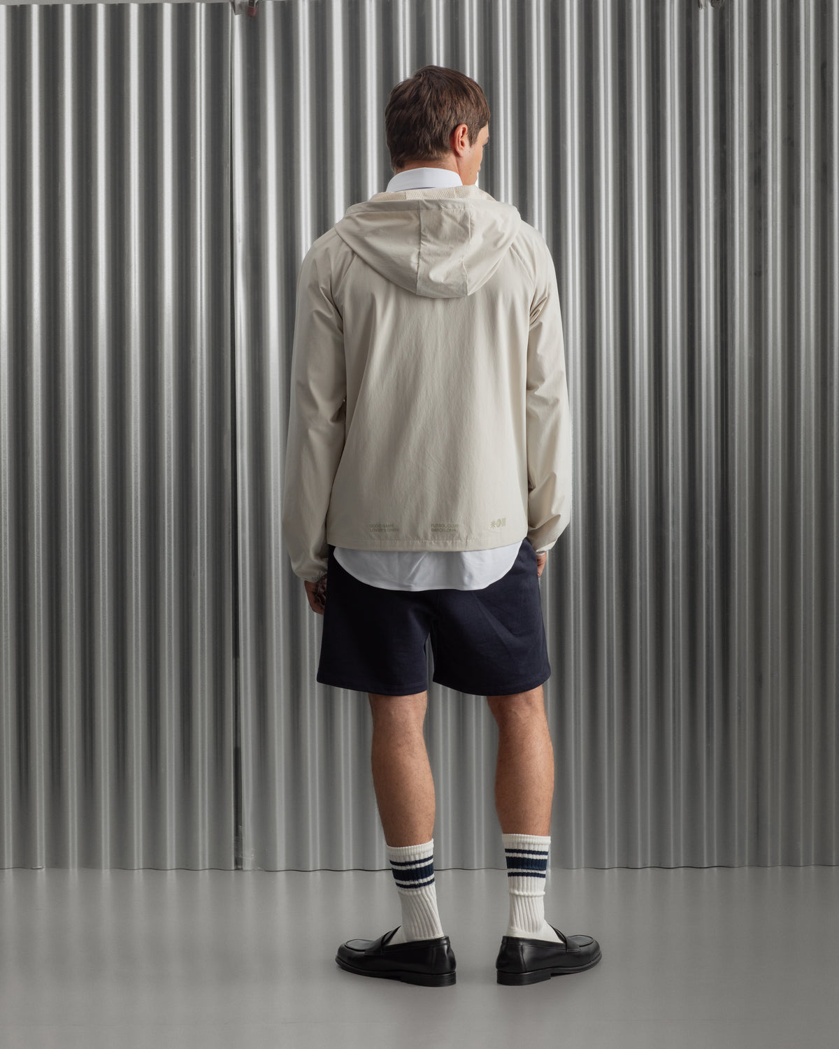 Man wearing a light beige FC Barcelona hooded jacket with front zipper, paired with navy shorts, white shirt, and striped socks, standing against a metallic background.