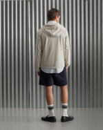 Man wearing a light beige FC Barcelona hooded jacket with front zipper, paired with navy shorts, white shirt, and striped socks, standing against a metallic background.