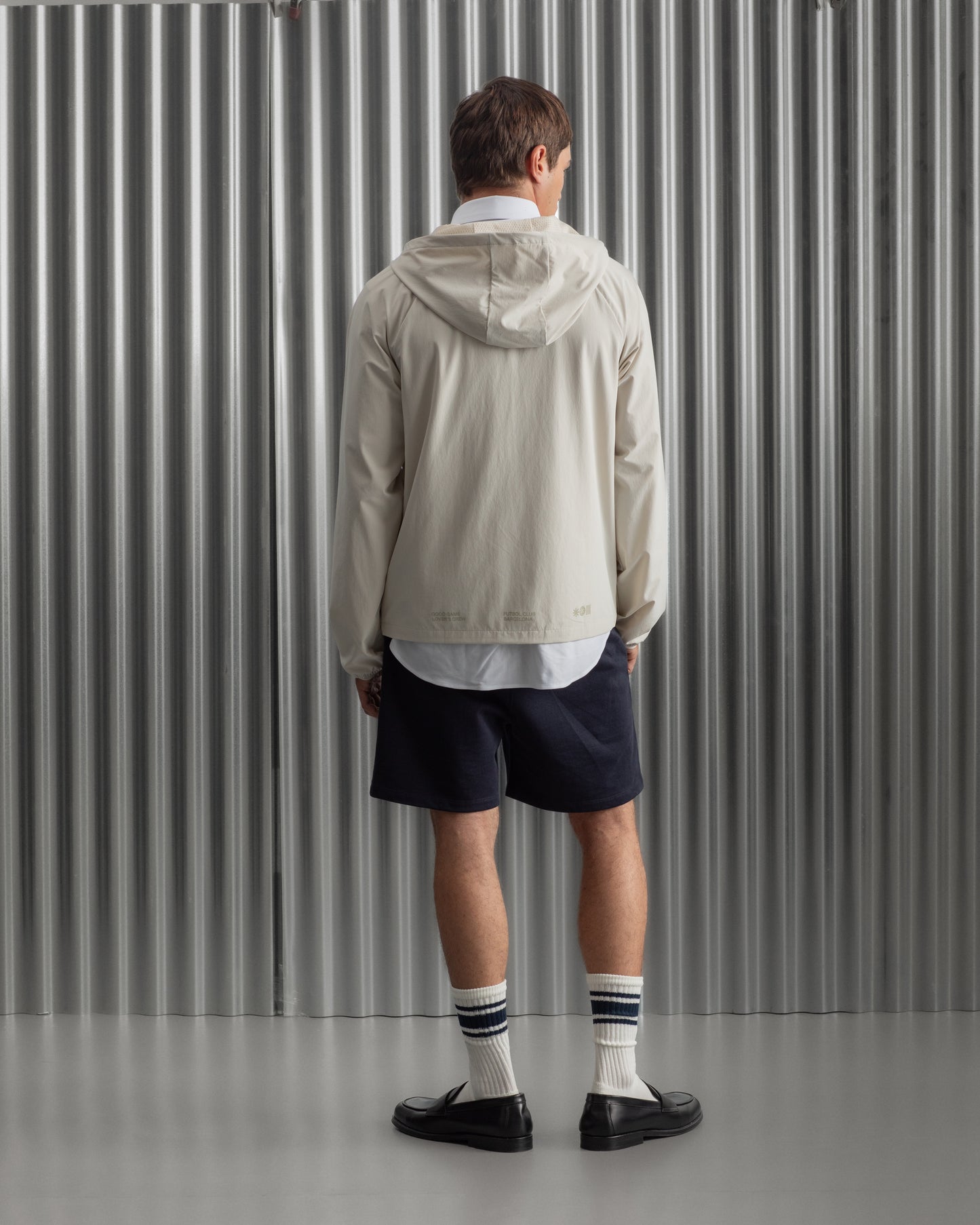 Man wearing a light beige FC Barcelona hooded jacket with front zipper, paired with navy shorts, white shirt, and striped socks, standing against a metallic background.