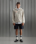 Man wearing a light beige FC Barcelona hooded jacket with front zipper, paired with navy shorts, white shirt, and striped socks, standing against a metallic background.