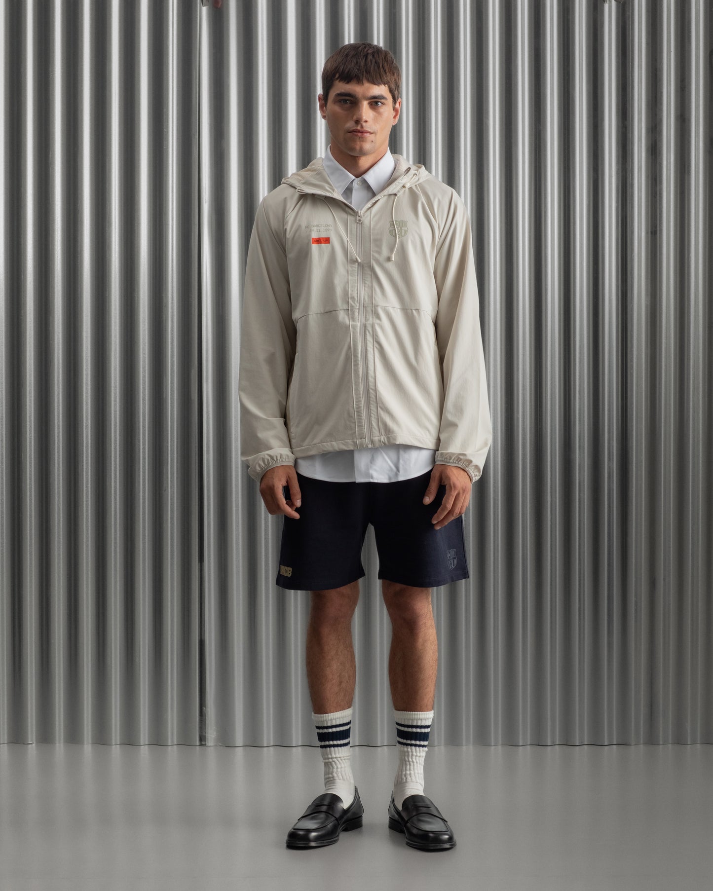 Man wearing a light beige FC Barcelona hooded jacket with front zipper, paired with navy shorts, white shirt, and striped socks, standing against a metallic background.