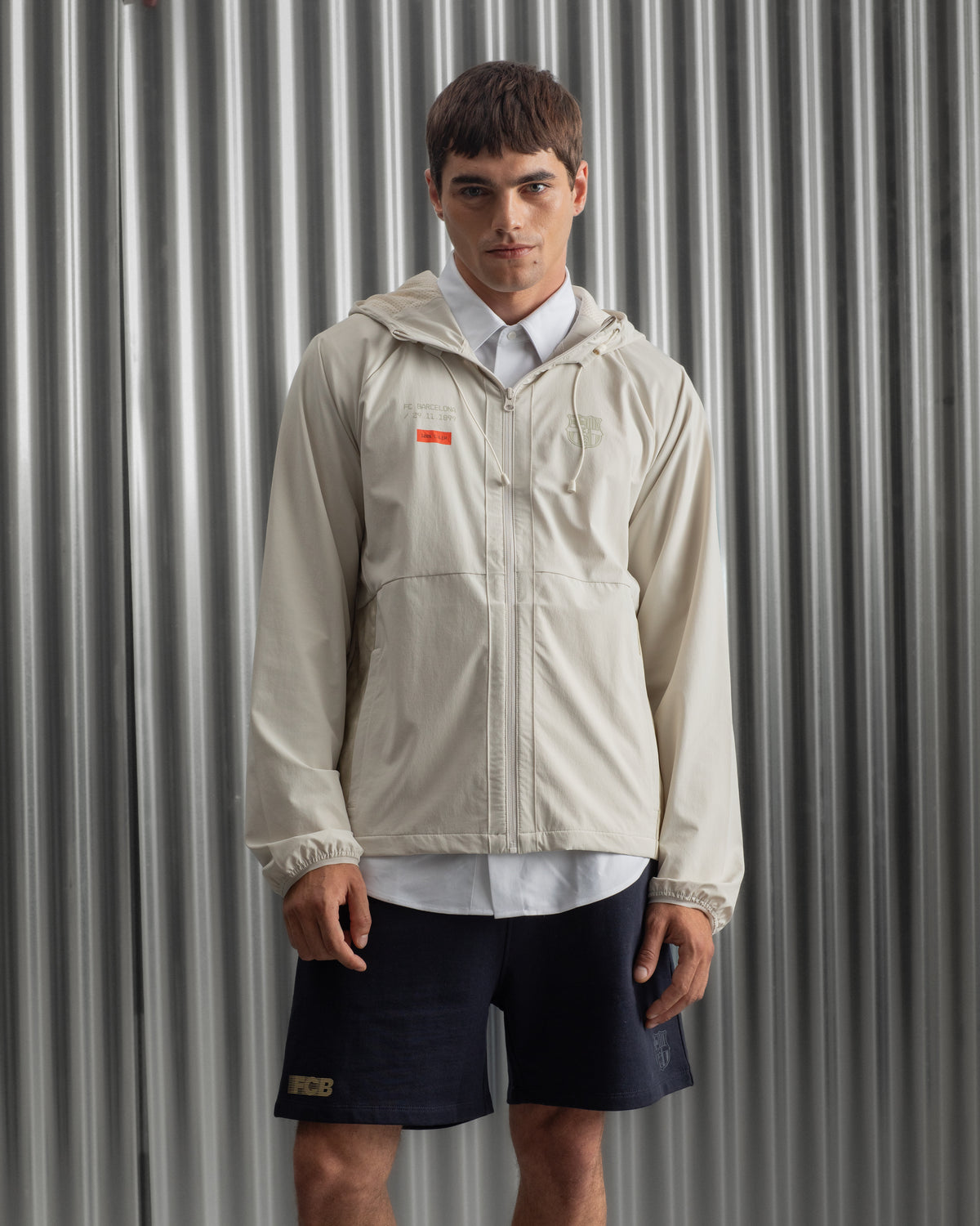 Man wearing a light beige FC Barcelona hooded jacket with front zipper, paired with navy shorts, white shirt, and striped socks, standing against a metallic background.
