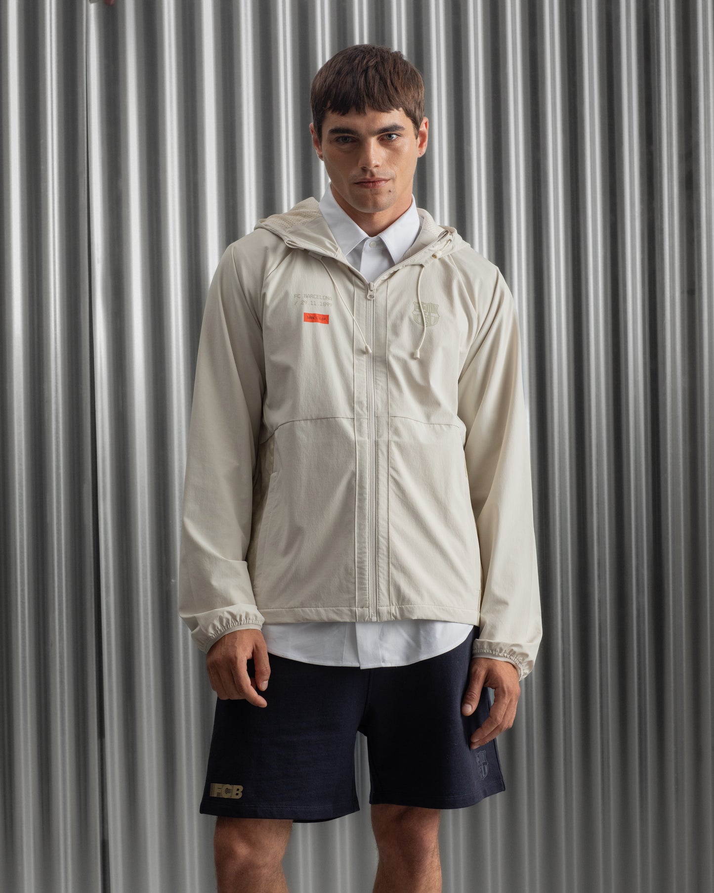 Man wearing a light beige FC Barcelona hooded jacket with front zipper, paired with navy shorts, white shirt, and striped socks, standing against a metallic background.