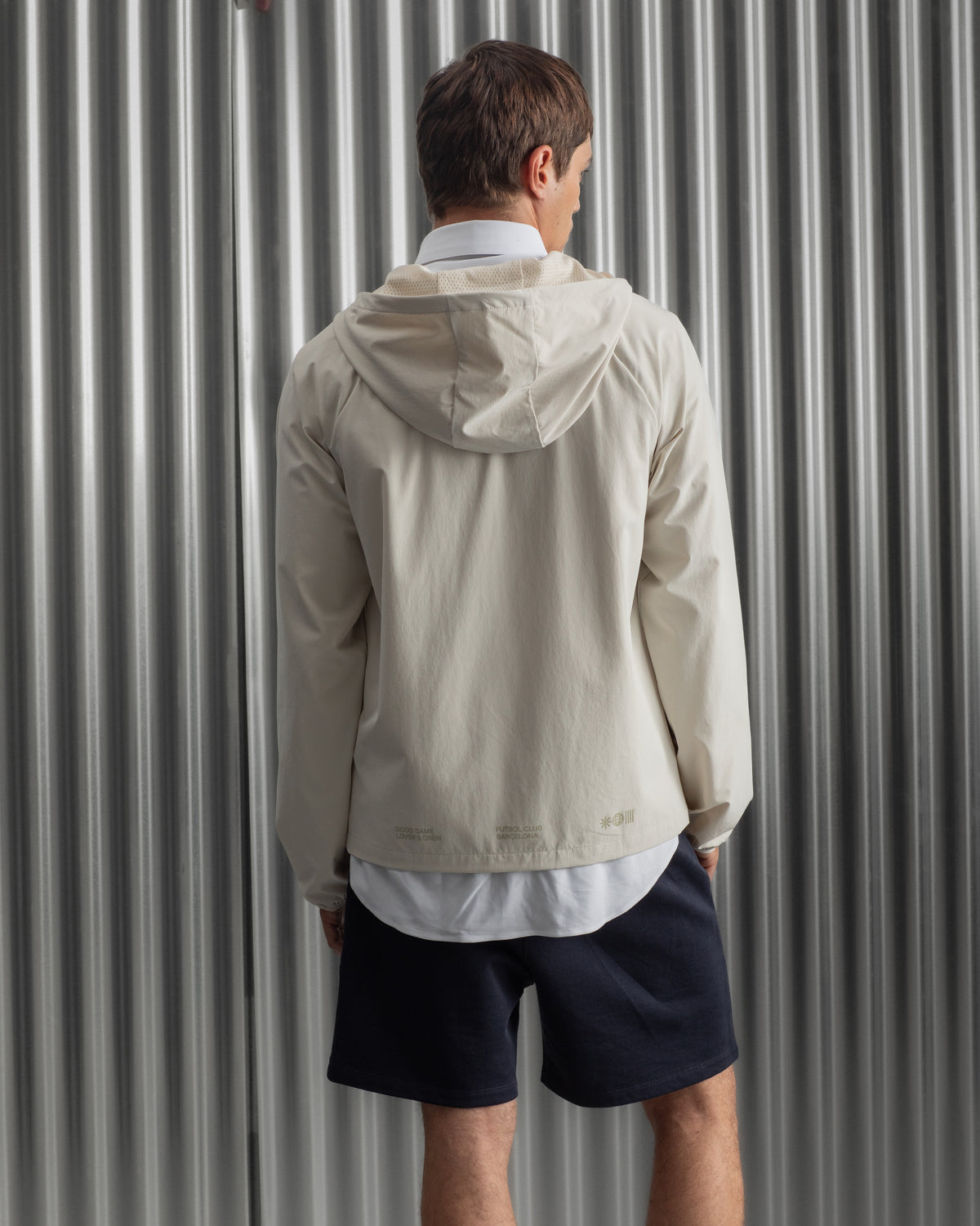 Man wearing a light beige FC Barcelona hooded jacket with front zipper, paired with navy shorts, white shirt, and striped socks, standing against a metallic background.