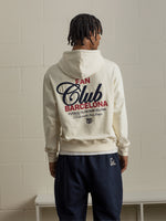 Man facing away from the camera wearing a cream hoodie with bold 'Fan Club Barcelona' text on the back, paired with navy shorts.