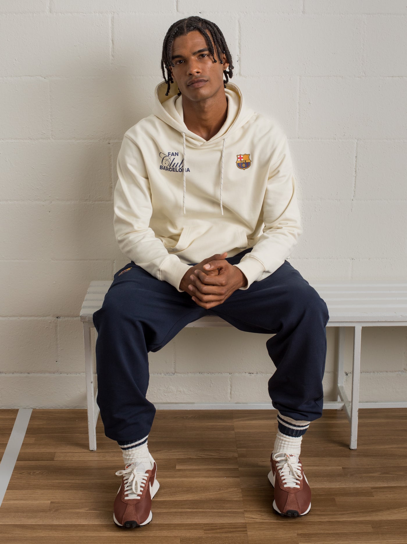 Man sitting on a white bench wearing a cream FC Barcelona hoodie with 'Fan Club Barcelona' text and navy sweatpants.
