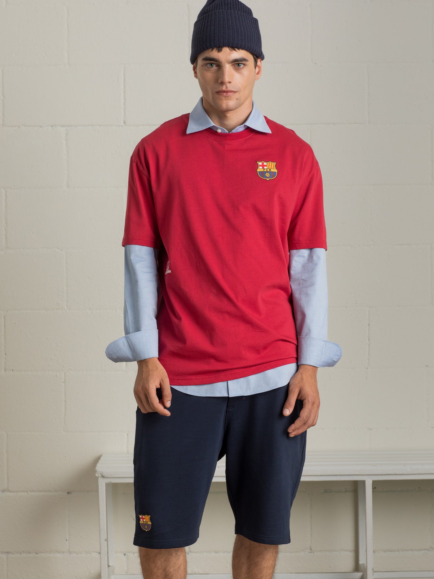 Man wearing a red 'Futbol Club Barcelona' T-shirt layered over a light blue shirt, navy shorts, and a dark beanie, facing the camera.