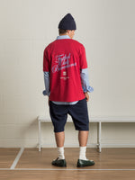 Man wearing a red 'Futbol Club Barcelona' T-shirt layered over a light blue shirt, navy shorts, and a dark beanie, facing away from the camera.